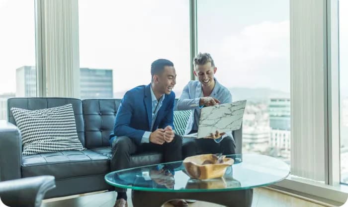 Two smiling businessmen in suits sitting on a couch, looking at a laptop and discussing a project in a modern office with a city view.