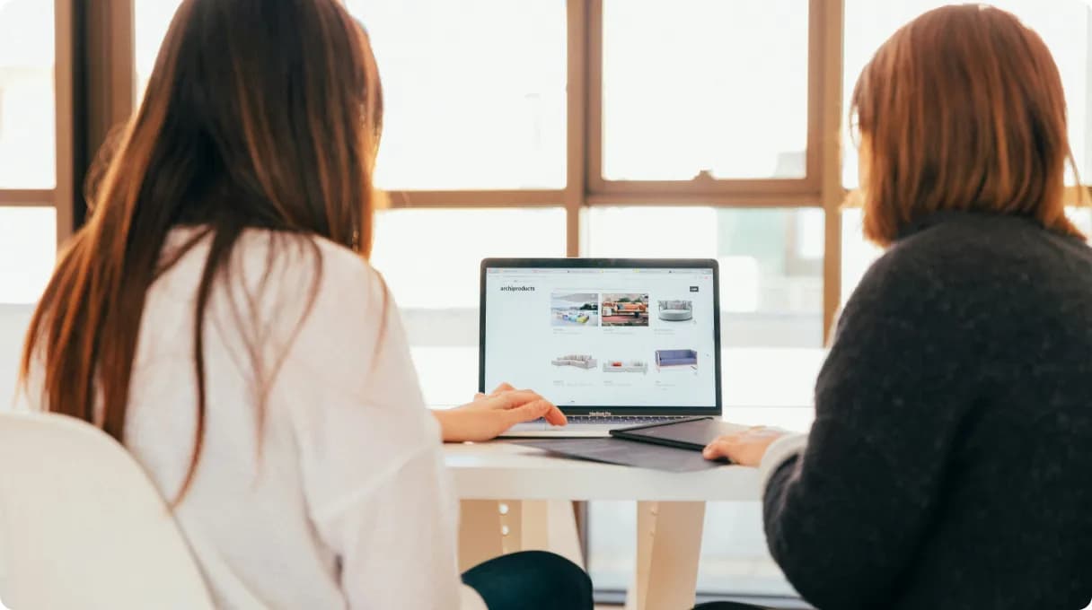 Two women sitting at a table with a laptop, discussing work together.